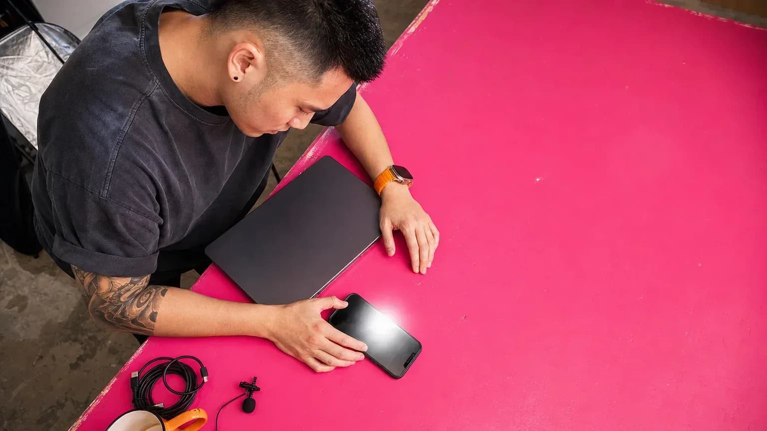 Seen from overhead, a creator leans over a magenta worktable thumbing a search into his phone beside a closed laptop