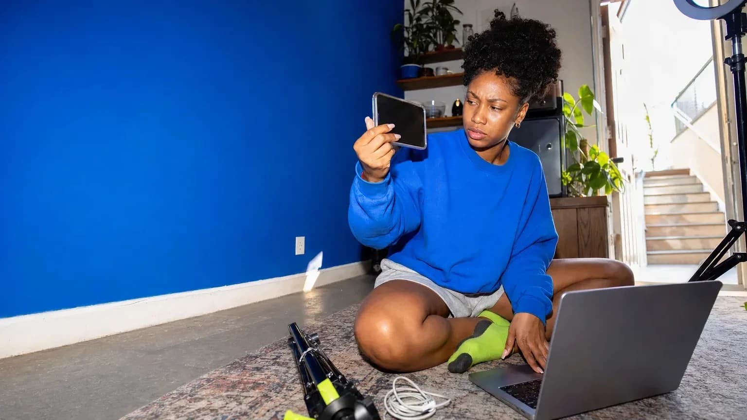 A Gen Z creator sits on her studio floor holding a phone up beside a laptop, comparing a reference video with her own remake