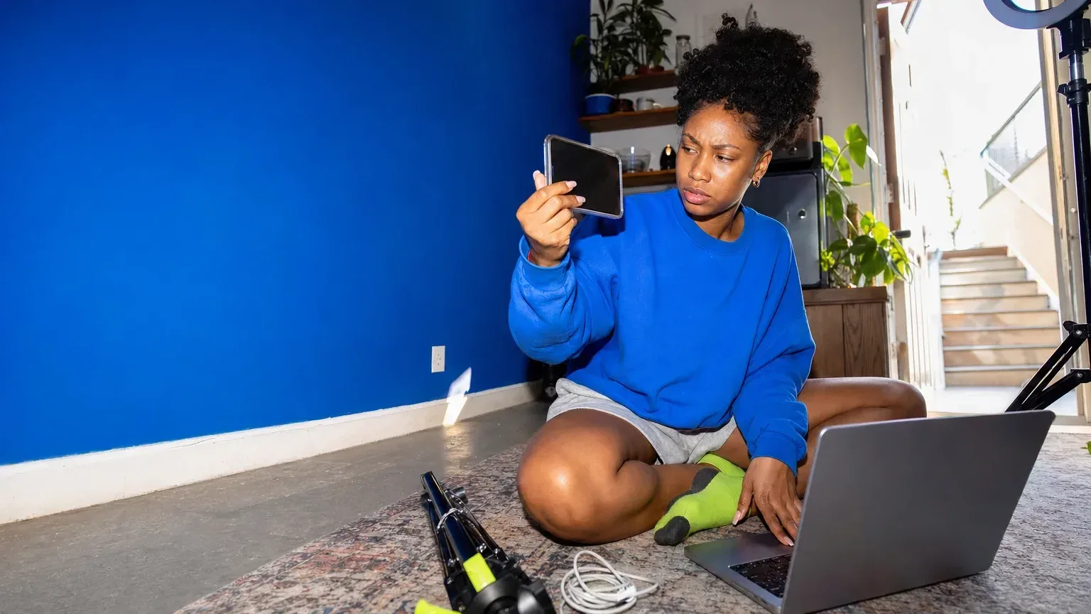 A Gen Z creator sits on her studio floor holding a phone up beside a laptop, comparing a reference video with her own remake